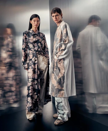 Two models wearing floral patterned matching outfits standing indoors against a metallic reflective backdrop in a fashion setting.
