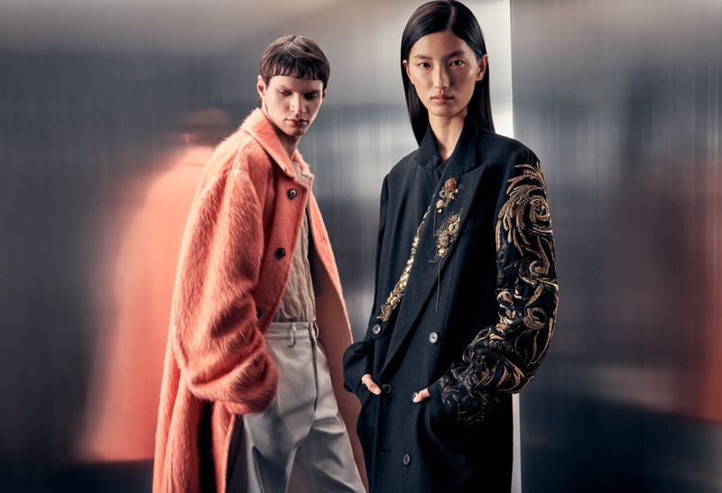Two people pose in a fashion editorial; a model in a pink faux-fur coat and light pants stands next to a model in a black embroidered coat.