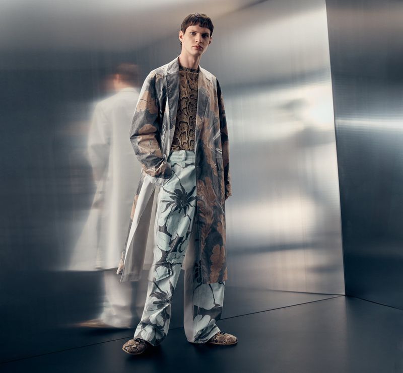 A young adult man poses in a patterned long coat and matching pants against a reflective wall, creating a fashion-forward editorial shot.