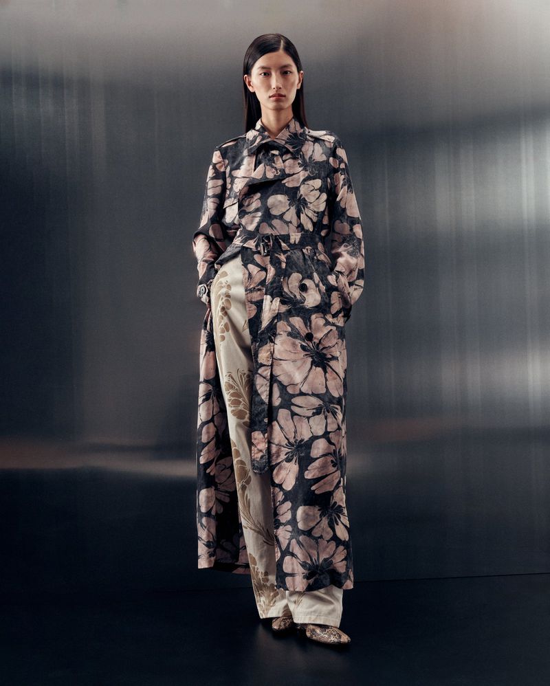 An Asian woman stands in a studio wearing a floral-print long coat and wide-leg pants with a light gray studio backdrop.