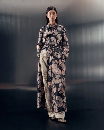An Asian woman stands in a studio wearing a floral-print long coat and wide-leg pants with a light gray studio backdrop.