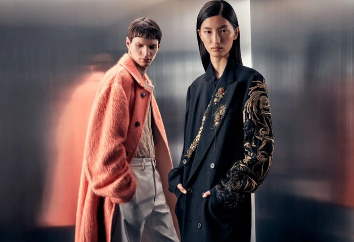 Two models posing against a metallic background, one wearing an orange textured coat and another in an embroidered black blazer.
