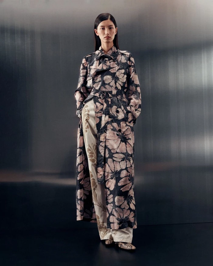 Model standing in a floral patterned trench coat and trousers against a dark brushed metallic background