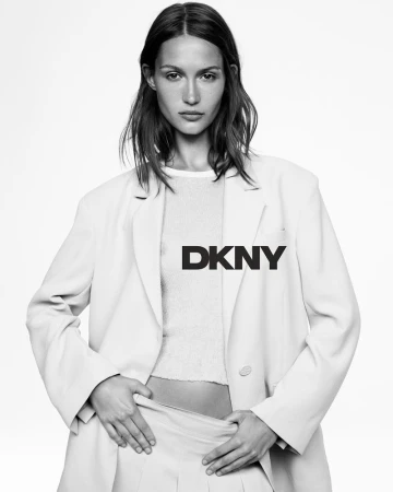 Black and white studio shot of an adult female fashion model wearing a blazer and skirt, logo overlay, posing against a white backdrop.