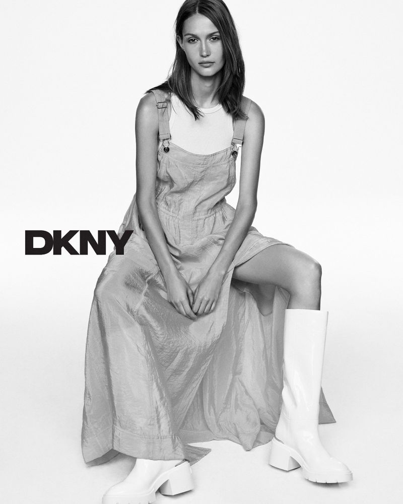 A woman in a gray sundress and white boots sits in a studio, highlighted in black and white. The DKNY logo appears on the left.