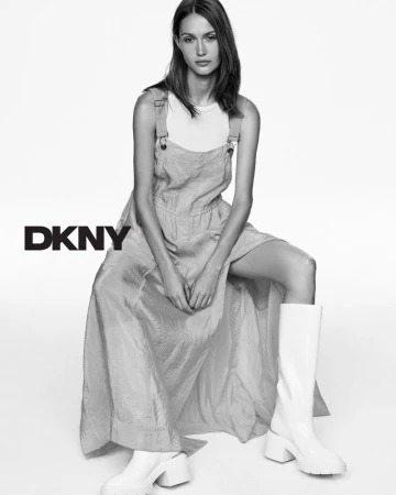 A woman in a gray sundress and white boots sits in a studio, highlighted in black and white. The DKNY logo appears on the left.