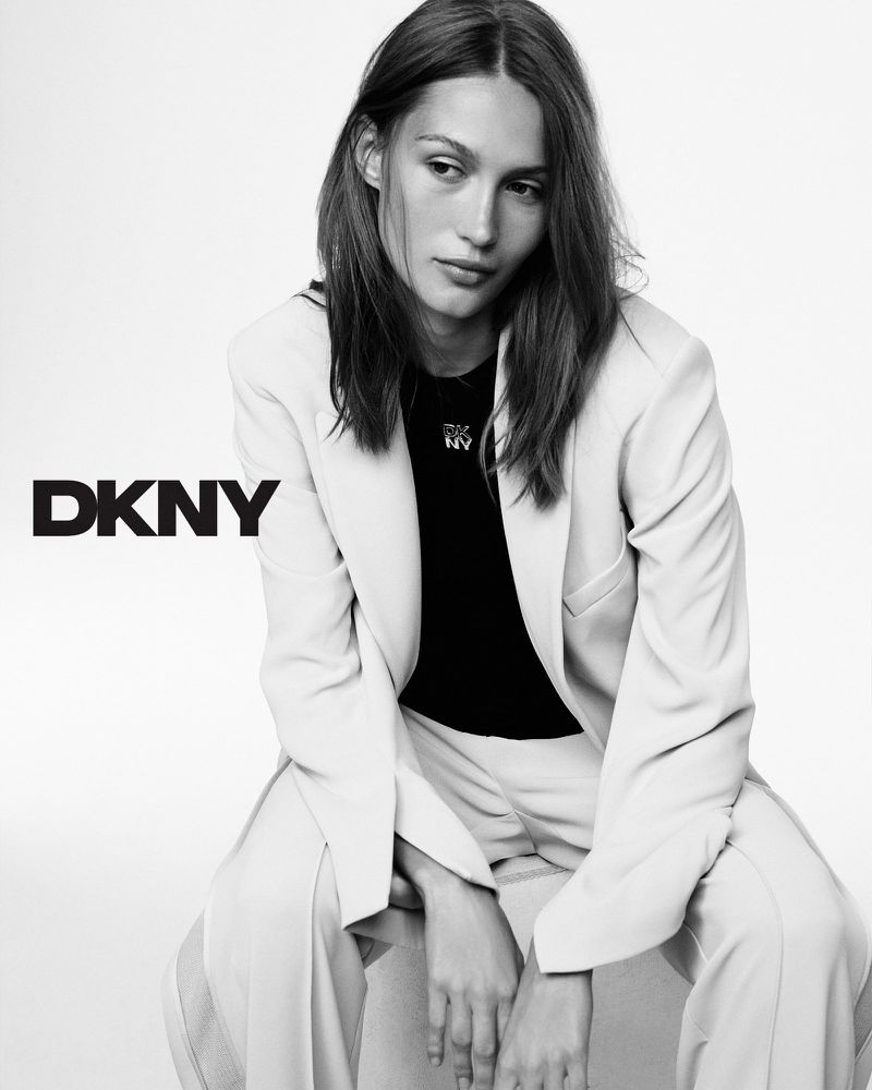 Black and white portrait of an adult woman in a blazer and pants sitting in a studio setting, promoting DKNY fashion.