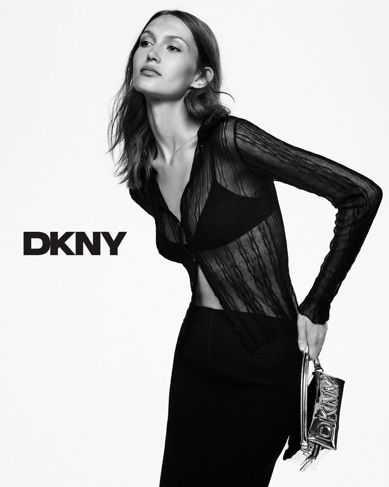 Black and white fashion shot of a woman in black top and skirt, holding a metallic DKNY purse against a bright, neutral background.