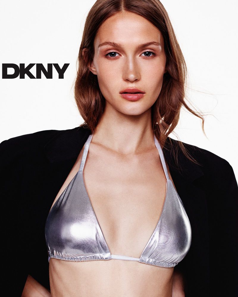 Model Vika Evseeva wearing a metallic silver halter top and black jacket against a plain white studio background for DKNY S/S 25