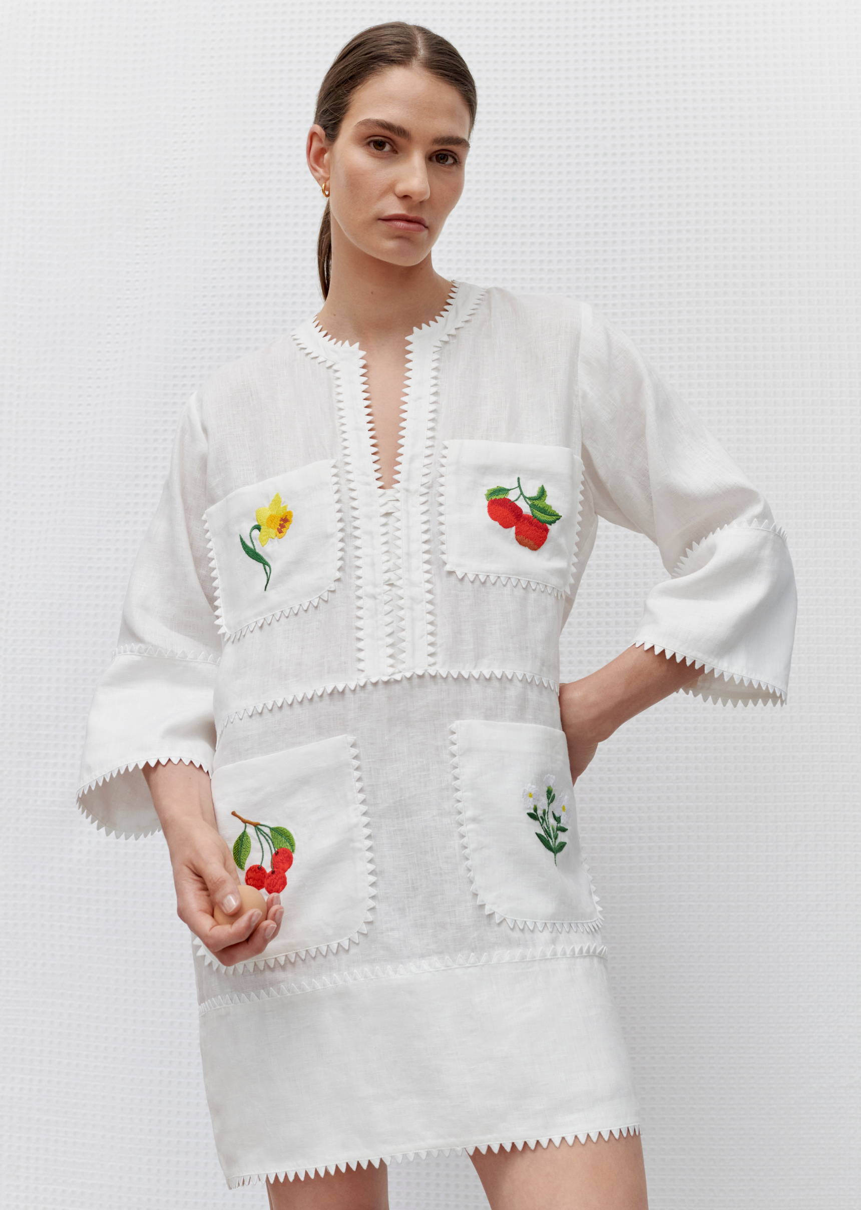 Adult woman wears a white mini dress with floral and cherry embroidery on the pockets, standing in a photo studio with neutral background.