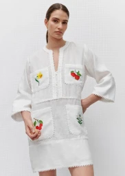 Model wearing a white embroidered linen dress with floral pocket details against a textured white background.