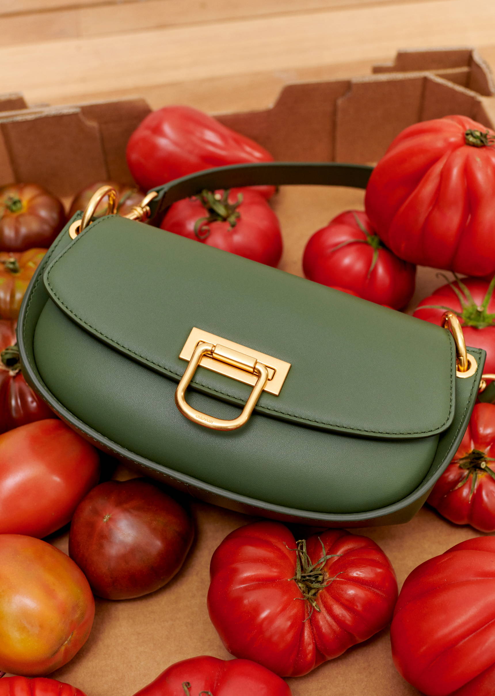 Olive green leather shoulder bag with gold hardware on a bed of vibrant red heirloom tomatoes and a cardboard background.