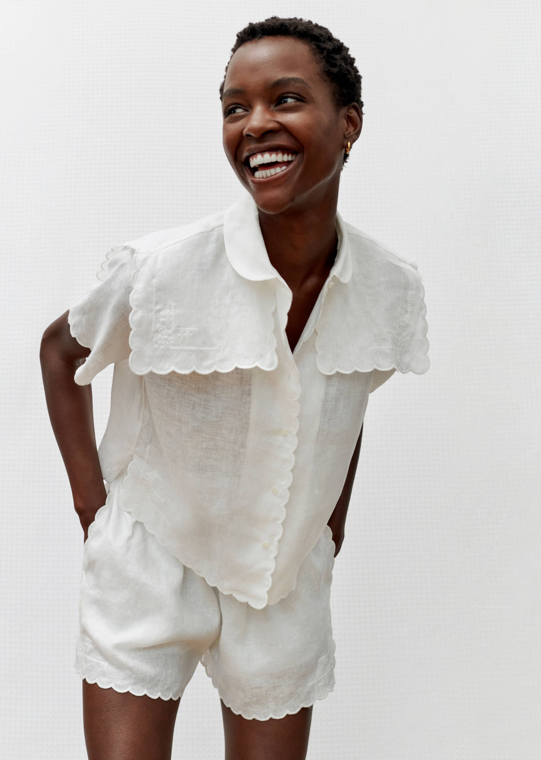 Smiling Black woman poses in a white linen short set with scalloped trim against a white background in a bright, soft light studio.