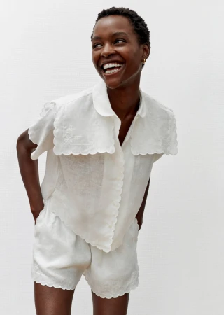 Smiling Black woman poses in a white linen short set with scalloped trim against a white background in a bright, soft light studio.