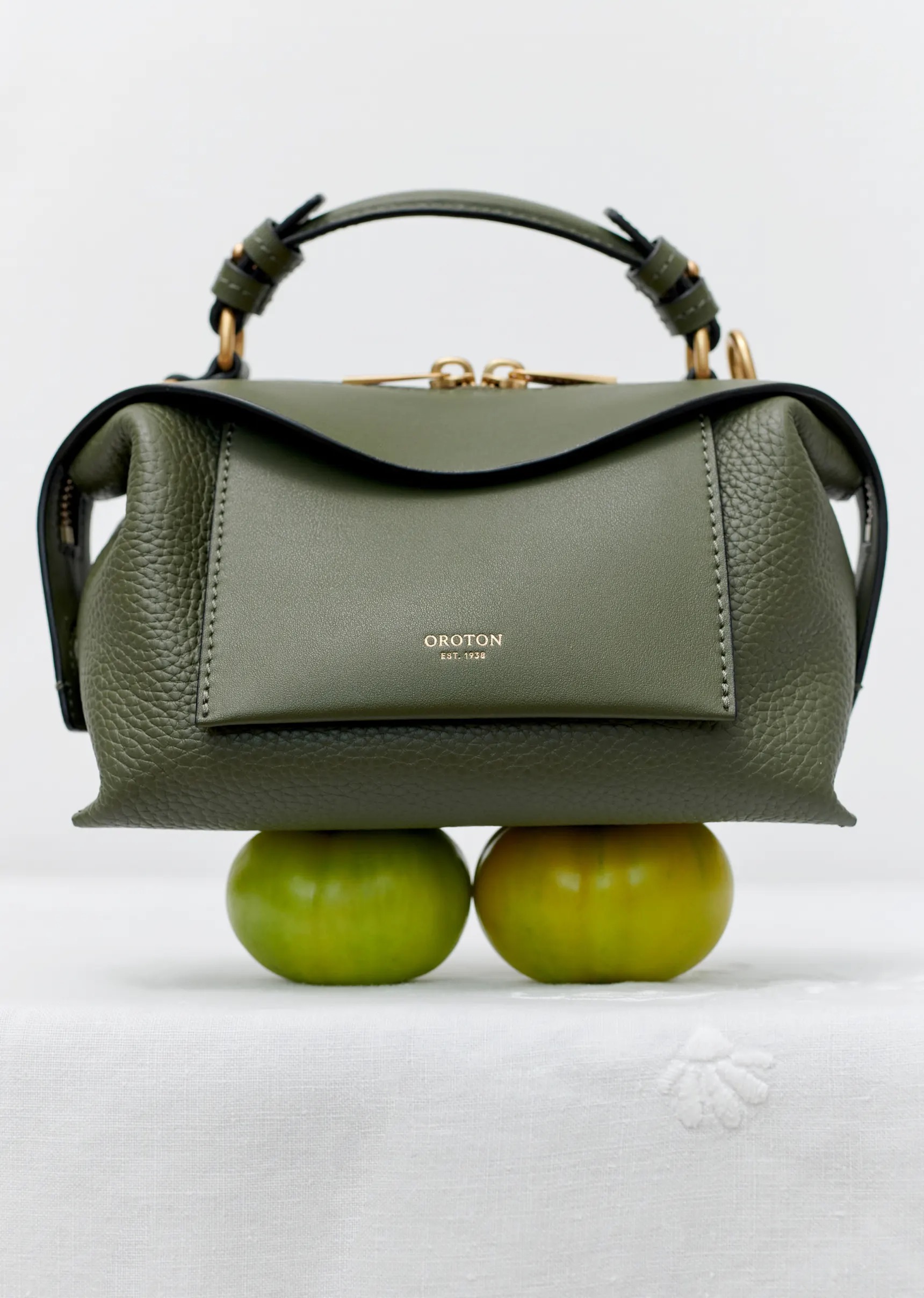 Olive green Oroton handbag with top handle and zip closure. The luxury purse sits atop two bright green apples on white fabric.