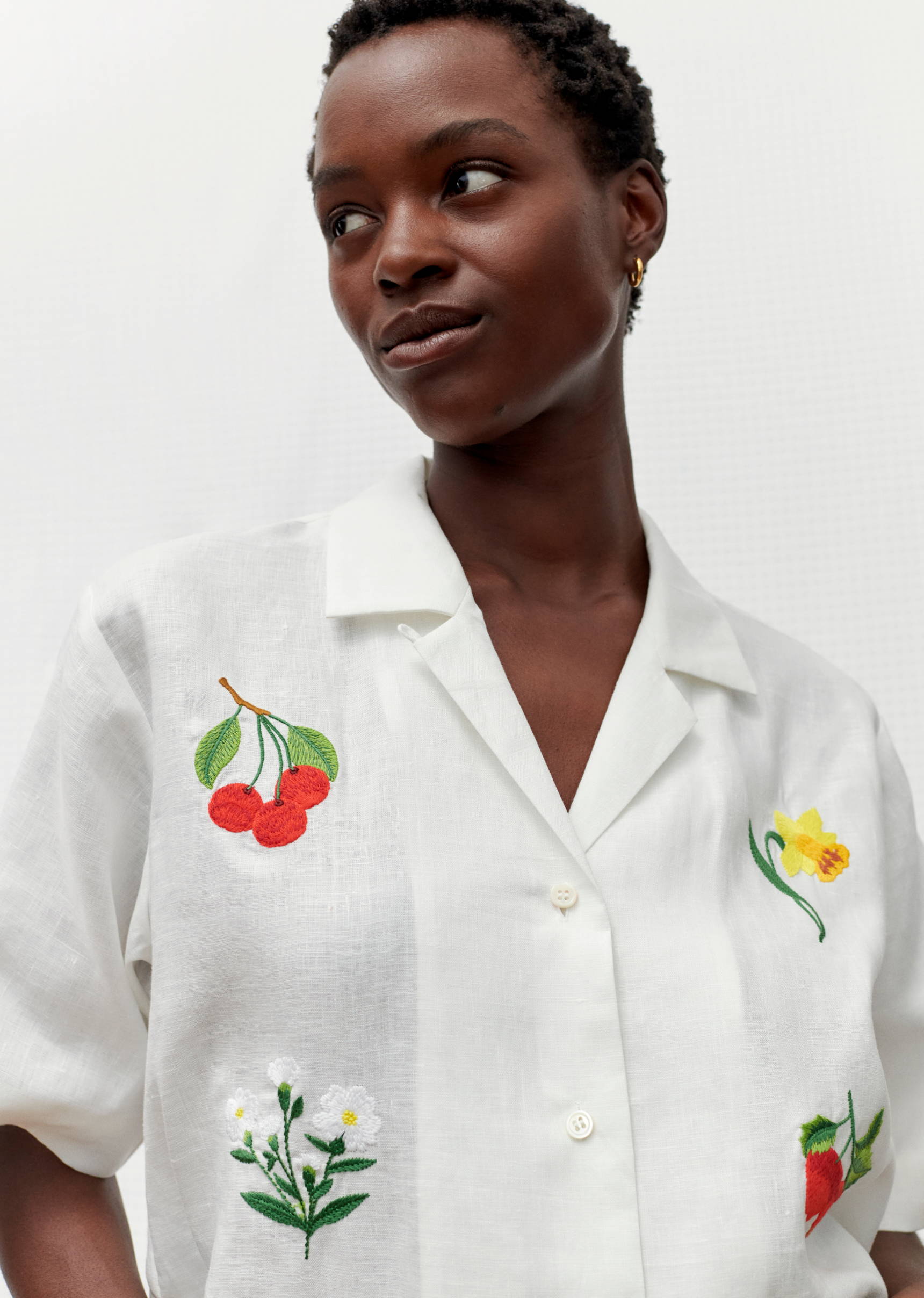 Woman wearing a white button-down shirt with floral and fruit embroidery, posing in front of a white wall in a bright setting.