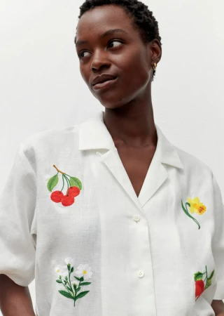 Woman wearing a white button-down shirt with floral and fruit embroidery, posing in front of a white wall in a bright setting.