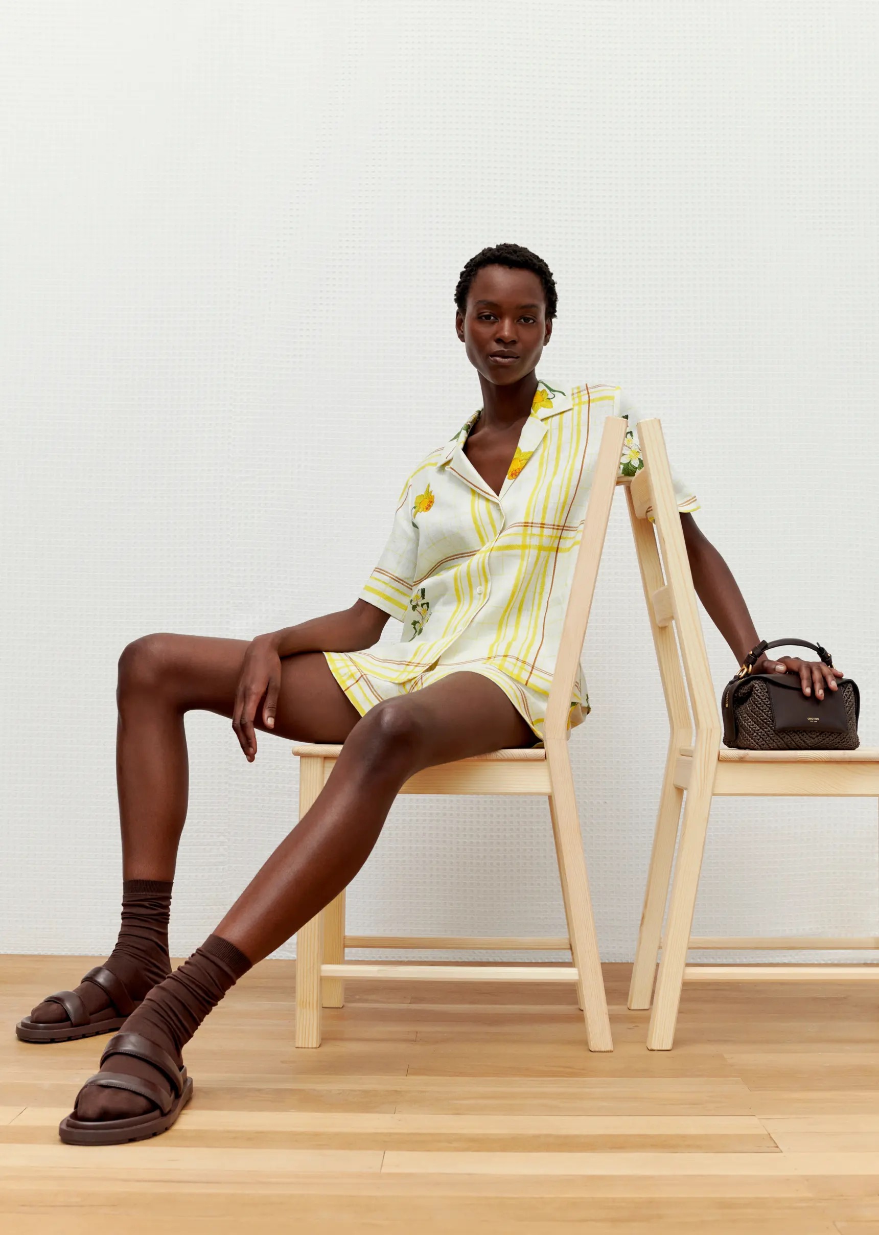 Dark-skinned model in yellow striped short set sits on wood chair with a brown handbag, brown socks, and sandal shoes against a white wall.