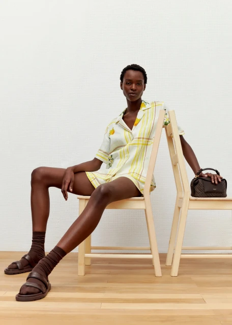 Dark-skinned model in yellow striped short set sits on wood chair with a brown handbag, brown socks, and sandal shoes against a white wall.