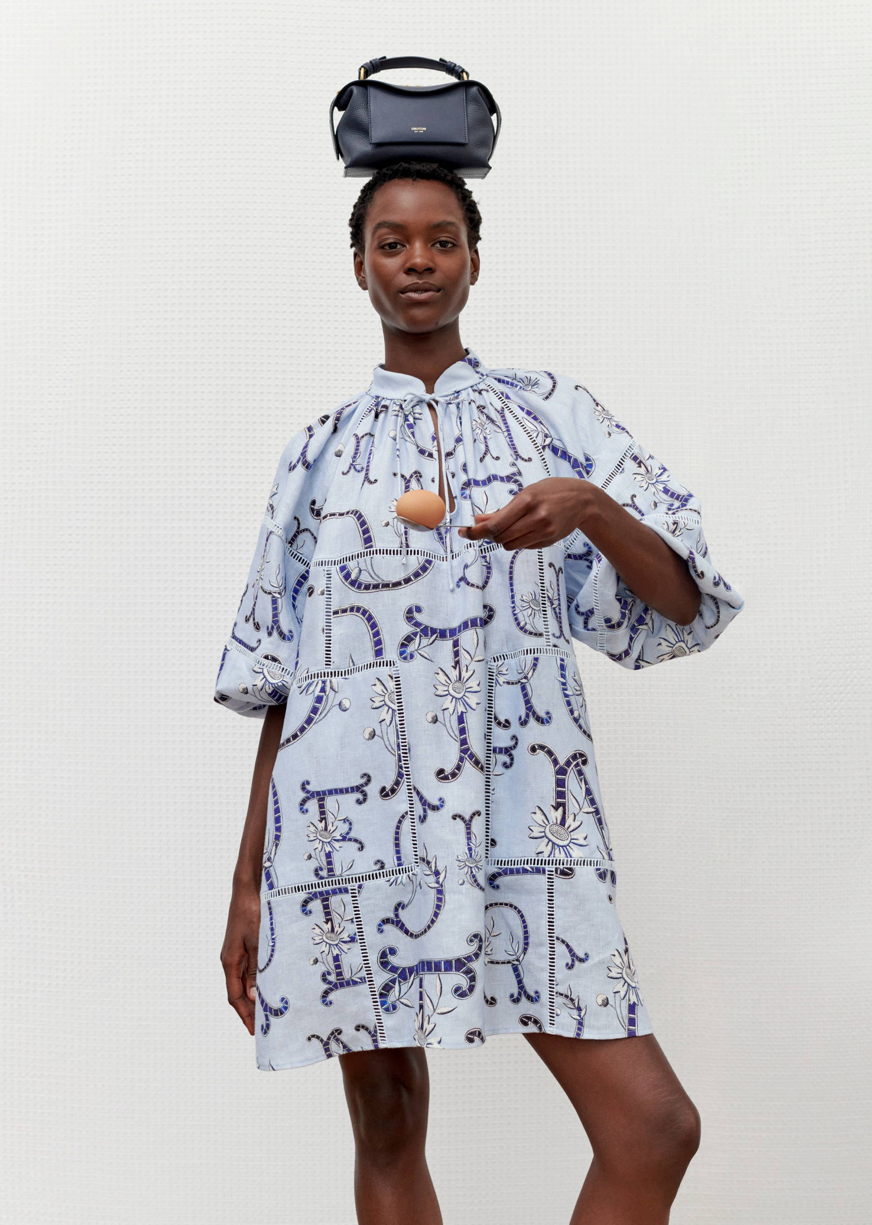 Adult woman balances a handbag on her head and an egg in her outstretched hand, wearing a patterned light blue dress.