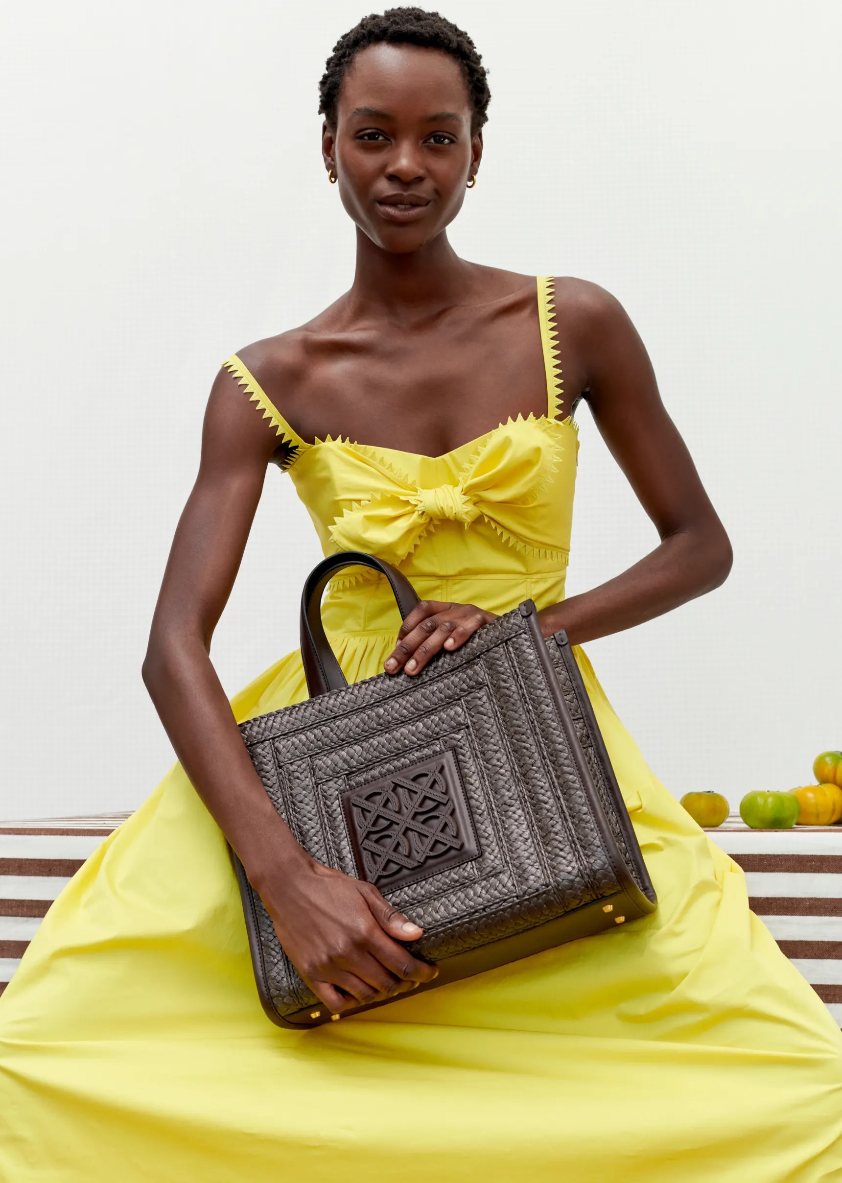 A black female fashion model in a yellow dress sits while holding a woven tote bag against a bright studio backdrop with fruit props.