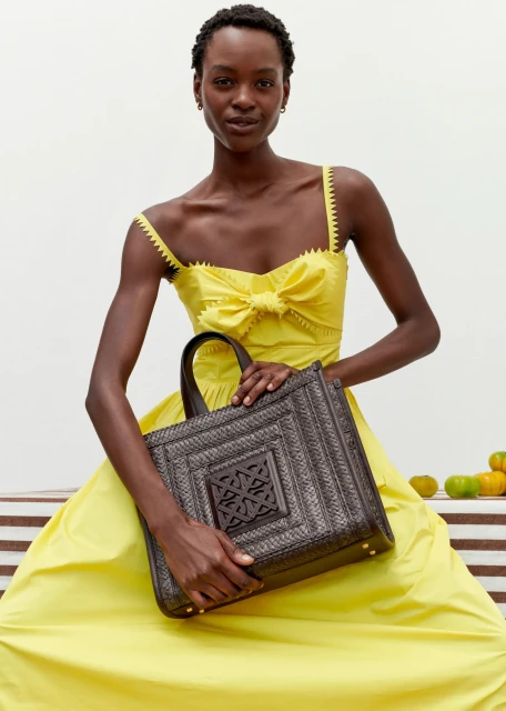 A black female fashion model in a yellow dress sits while holding a woven tote bag against a bright studio backdrop with fruit props.