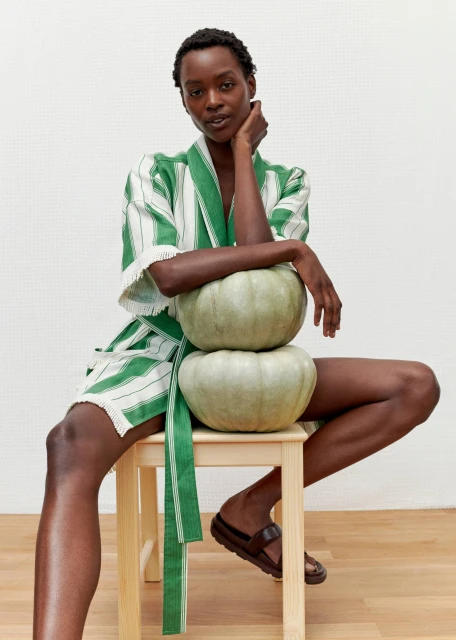 A slender black woman sits on a wooden stool, posing with stacked green pumpkins in a studio, wearing a striped robe and sandals.