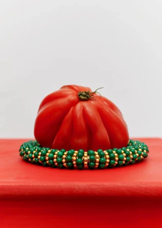 Ripe heirloom tomato on a beaded necklace, set on a red surface against a white background, simple composition, studio setting.