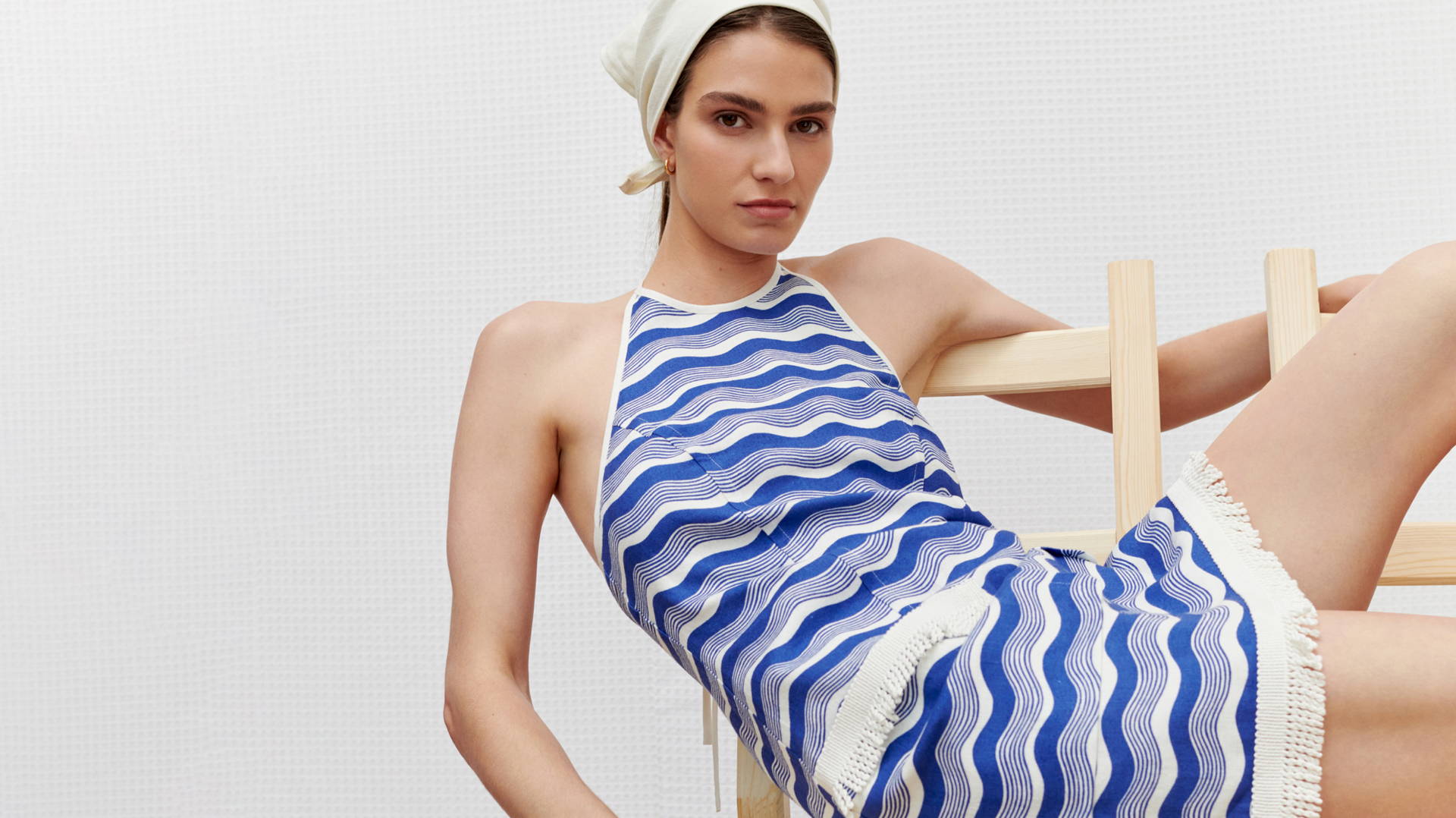 Brunette woman wearing a blue and white striped halter dress and headscarf posing on a light wooden chair against a white background.