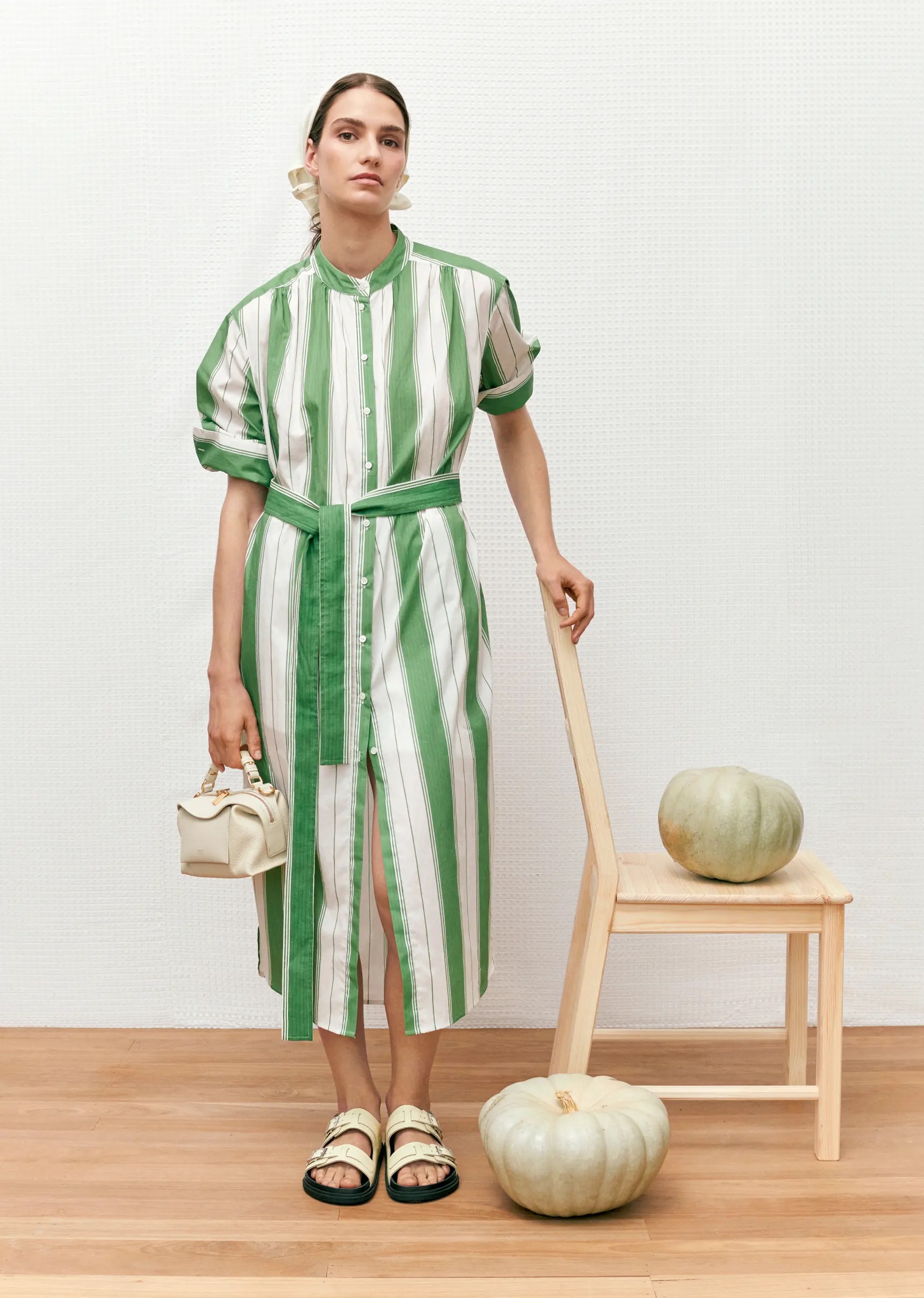 A fair-skinned woman models a striped green and white dress with white sandals, accessorized with white earrings and handbag, posing indoors.
