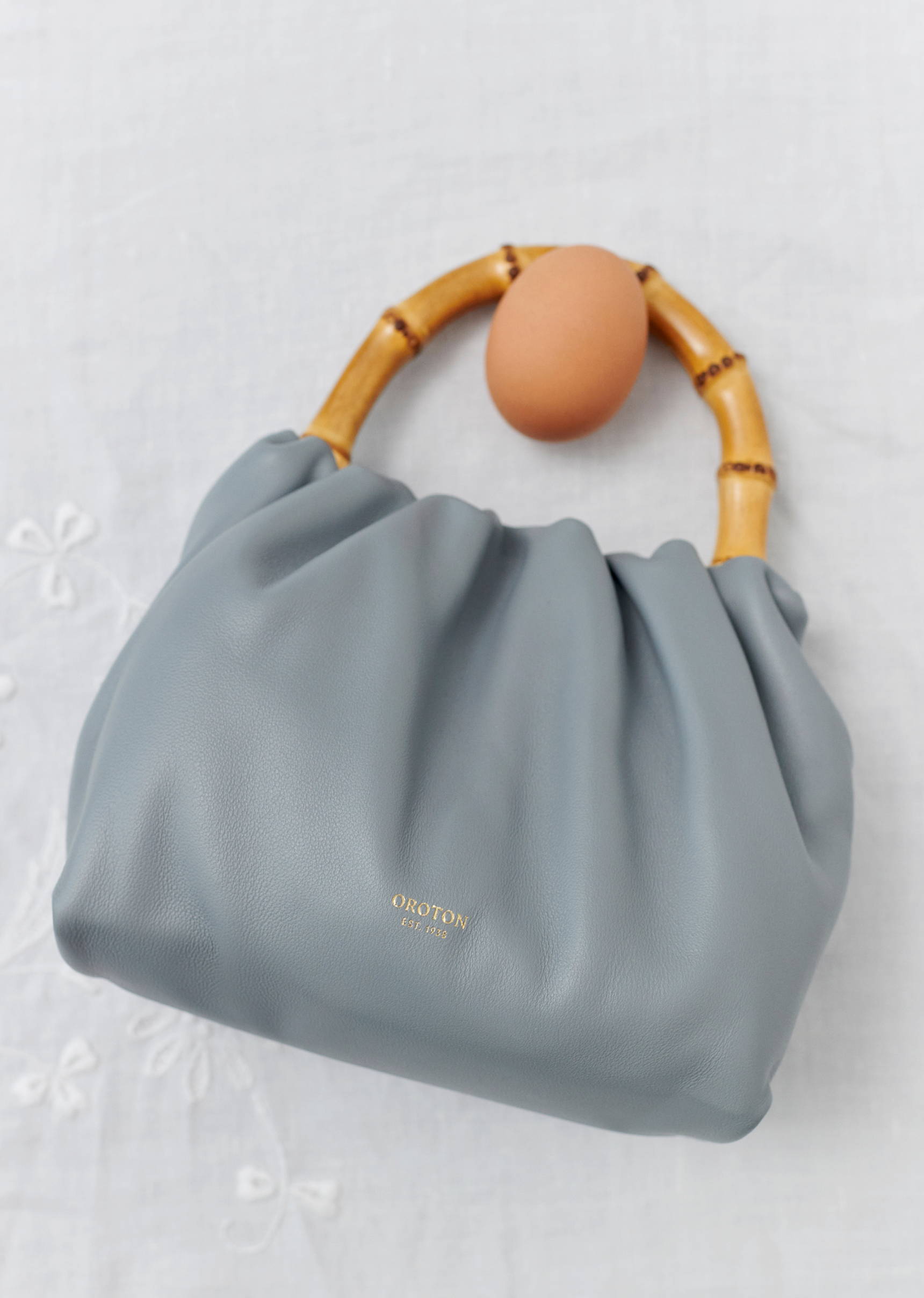 Light gray Oroton handbag with a bamboo handle and an egg perched on top is set against an off-white floral patterned surface.
