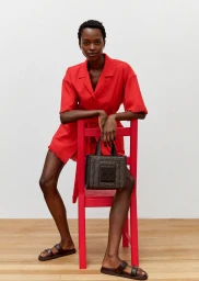 Model in a red dress leaning on a pink chair holding a dark woven handbag against a seamless white background.
