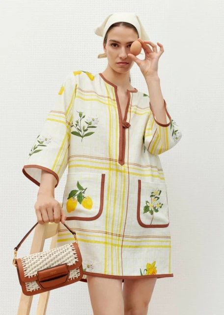 Woman in a white and yellow dress and headscarf holds an egg up to her eye, standing indoors with a brown purse on her arm.