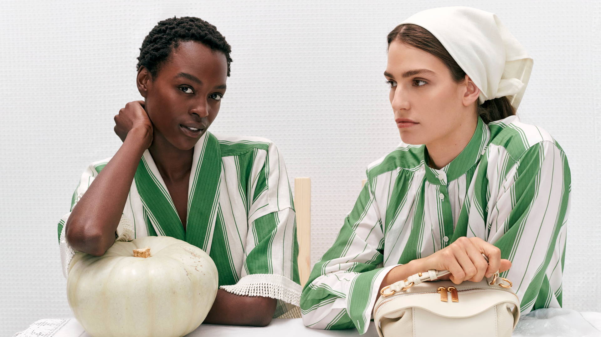 Two fashion models in striped pajamas pose indoors with a handbag and a pumpkin, showcasing beauty and fashion in a studio setting.