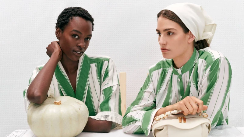 Two fashion models in striped pajamas pose indoors with a handbag and a pumpkin, showcasing beauty and fashion in a studio setting.