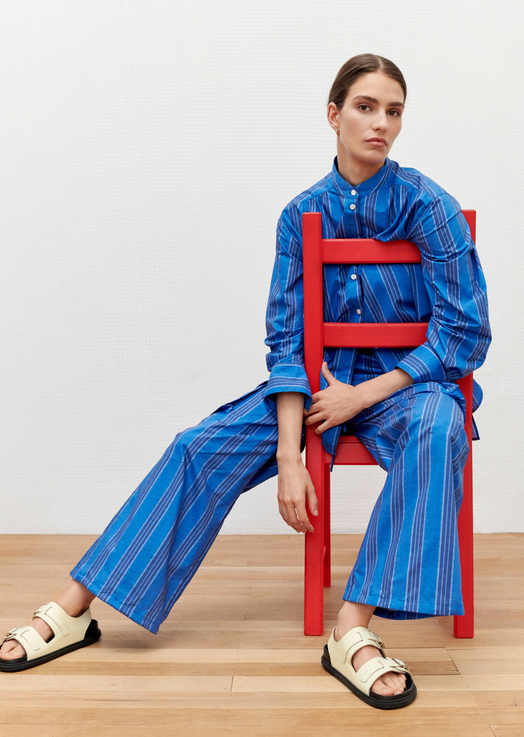 Fashion shot of a woman in a blue striped shirt and pants sitting on a red chair indoors against a white backdrop, wearing cream sandals.