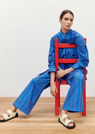 Fashion shot of a woman in a blue striped shirt and pants sitting on a red chair indoors against a white backdrop, wearing cream sandals.