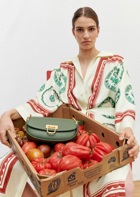 A woman in a patterned shirt dress sits with a green purse in a cardboard box full of red tomatoes indoors against a white wall.