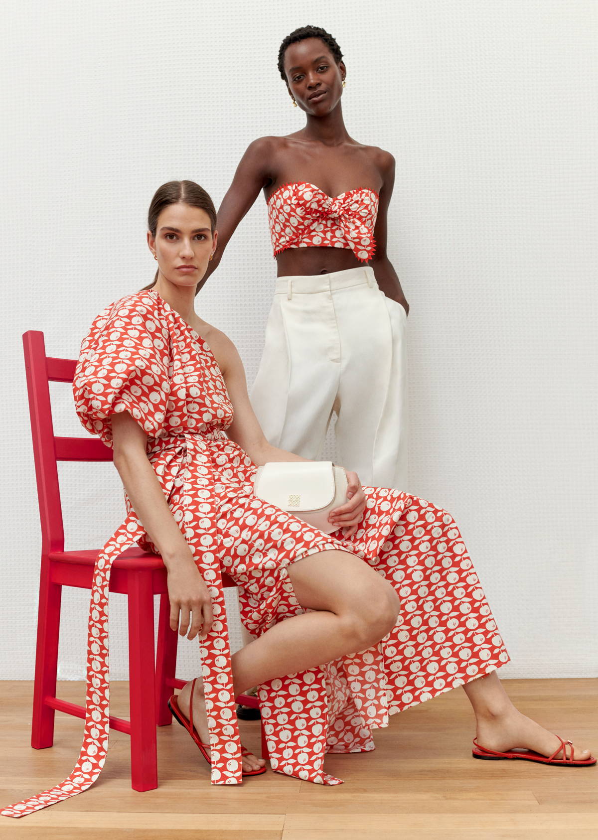 Two fashion models pose in patterned summer outfits against a white wall. One sits on a red chair holding a small white purse.