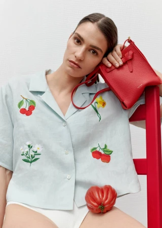 Model in pale blue embroidered shirt holding a red handbag while leaning on a red chair against a white background