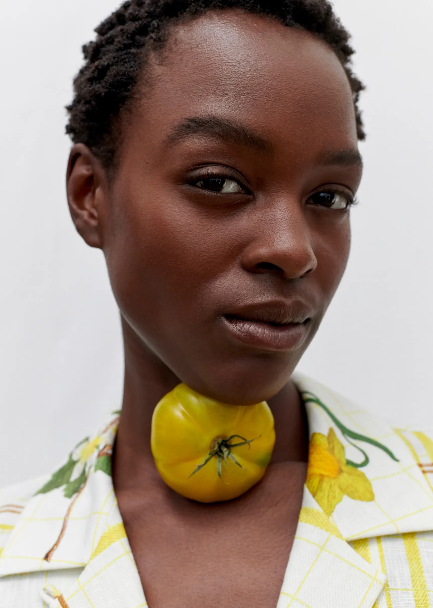 Close-up of a woman with short hair wearing a floral jacket and yellow tomato necklace against a white background.