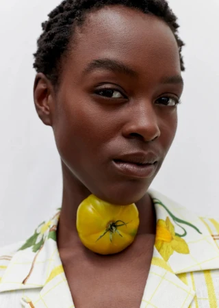 Close-up of a woman with short hair wearing a floral jacket and yellow tomato necklace against a white background.