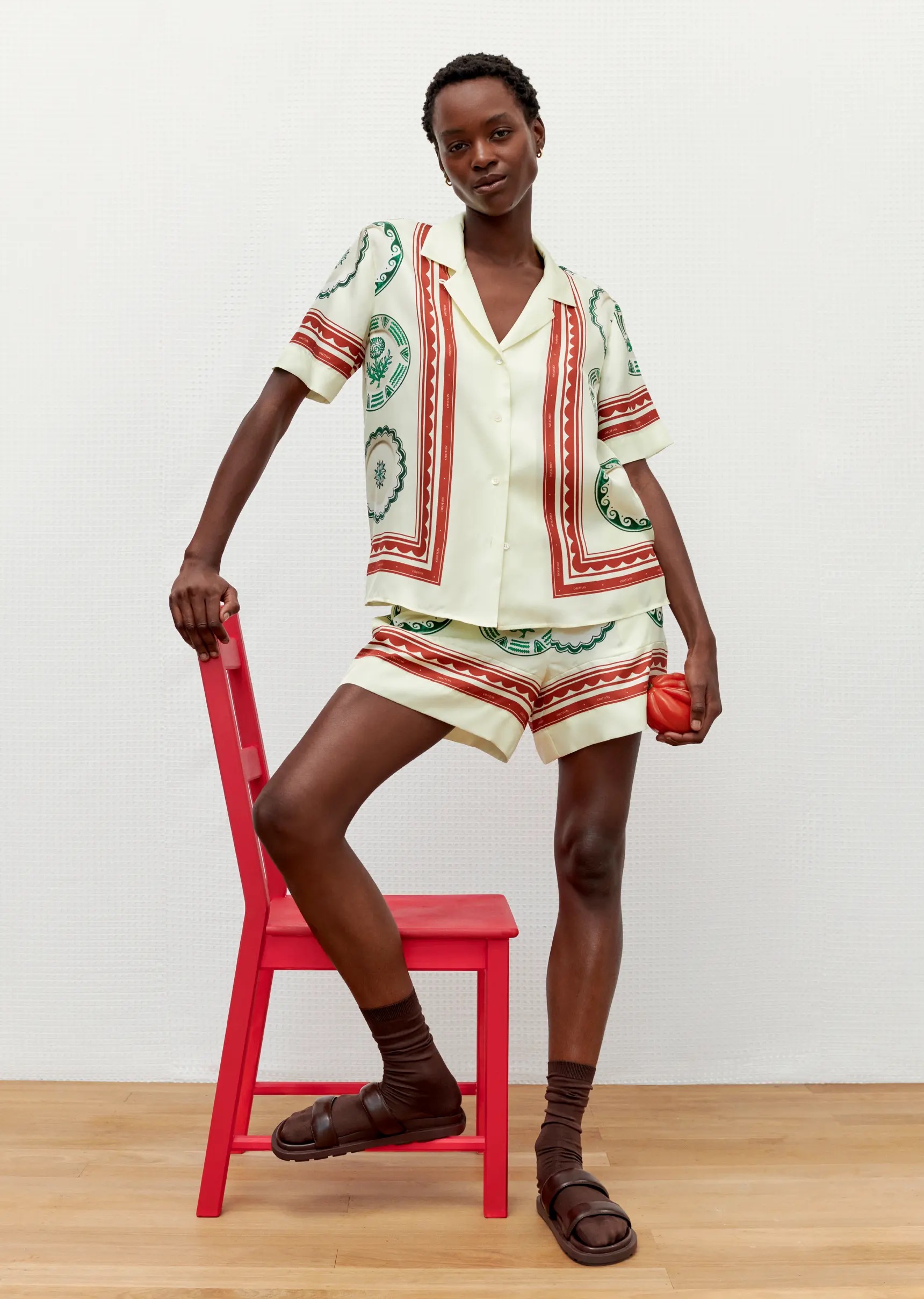 Woman posing in patterned short set with chair and sandals in a bright studio, showcasing contemporary fashion and simple studio photography.