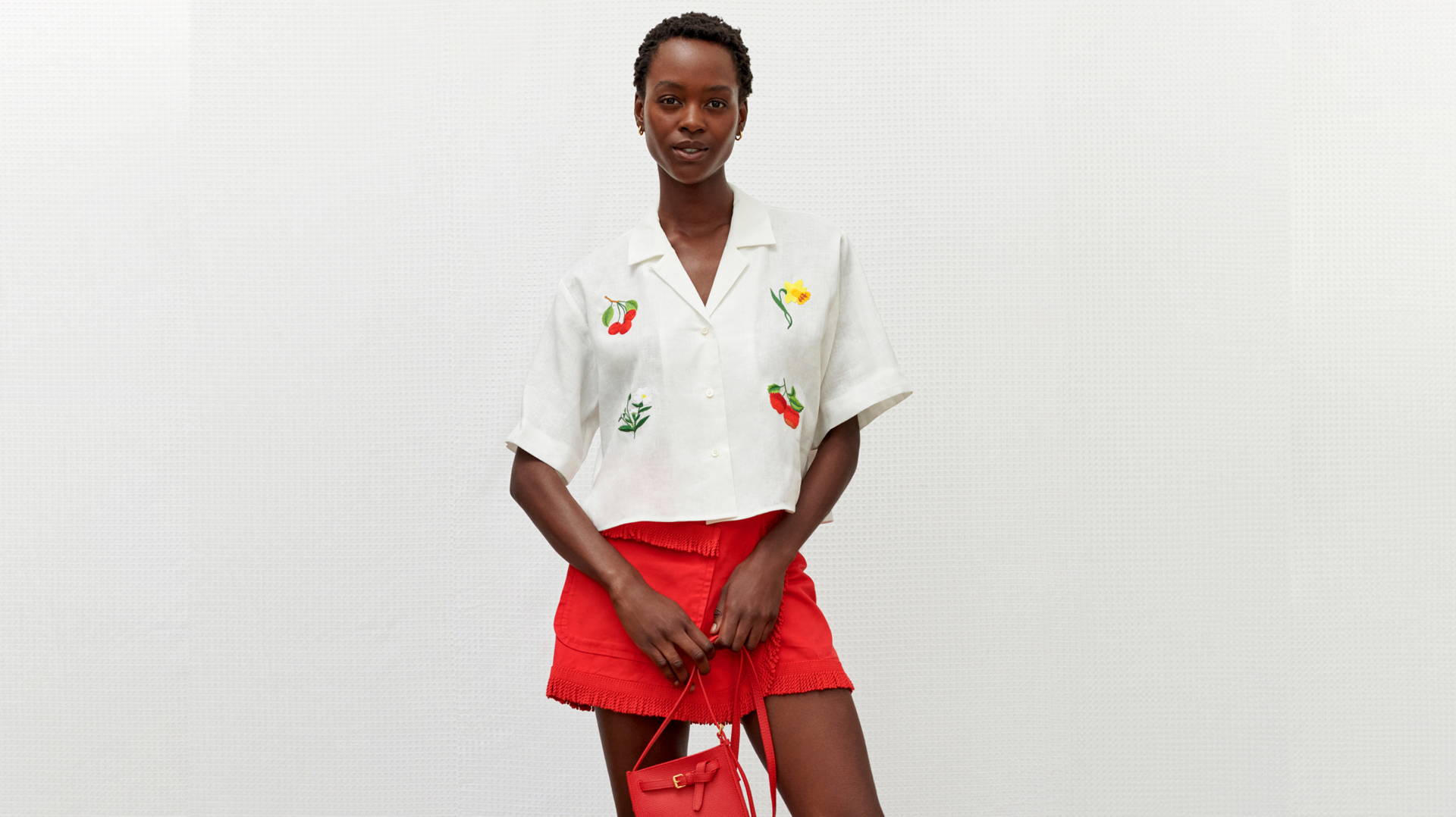 Model wearing a white embroidered short-sleeve shirt and a red skirt holding a matching red handbag against a textured backdrop