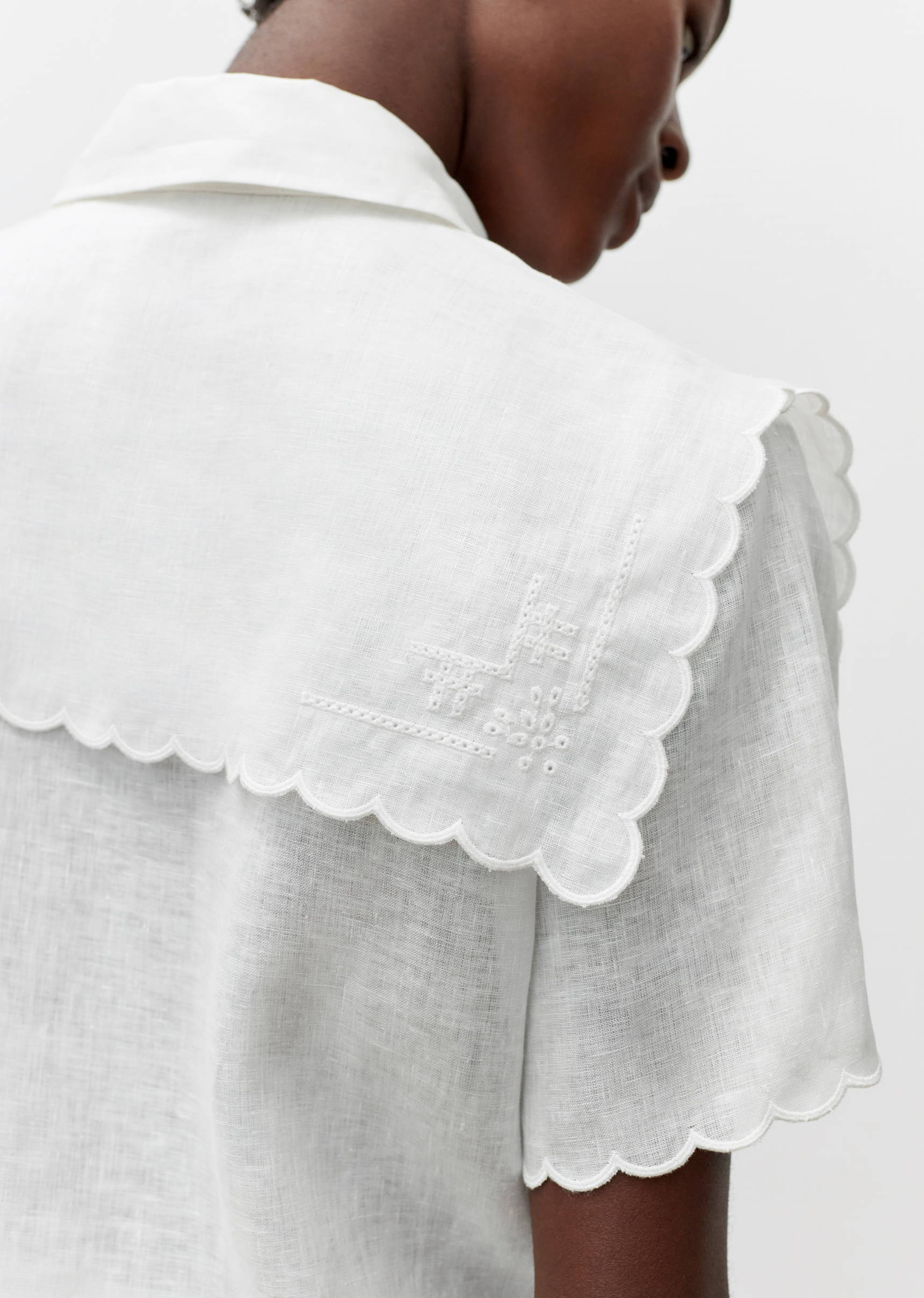 An adult female displays a short-sleeved white top with a large, embroidered collar and scalloped edges, shot with a focus on fashion details.