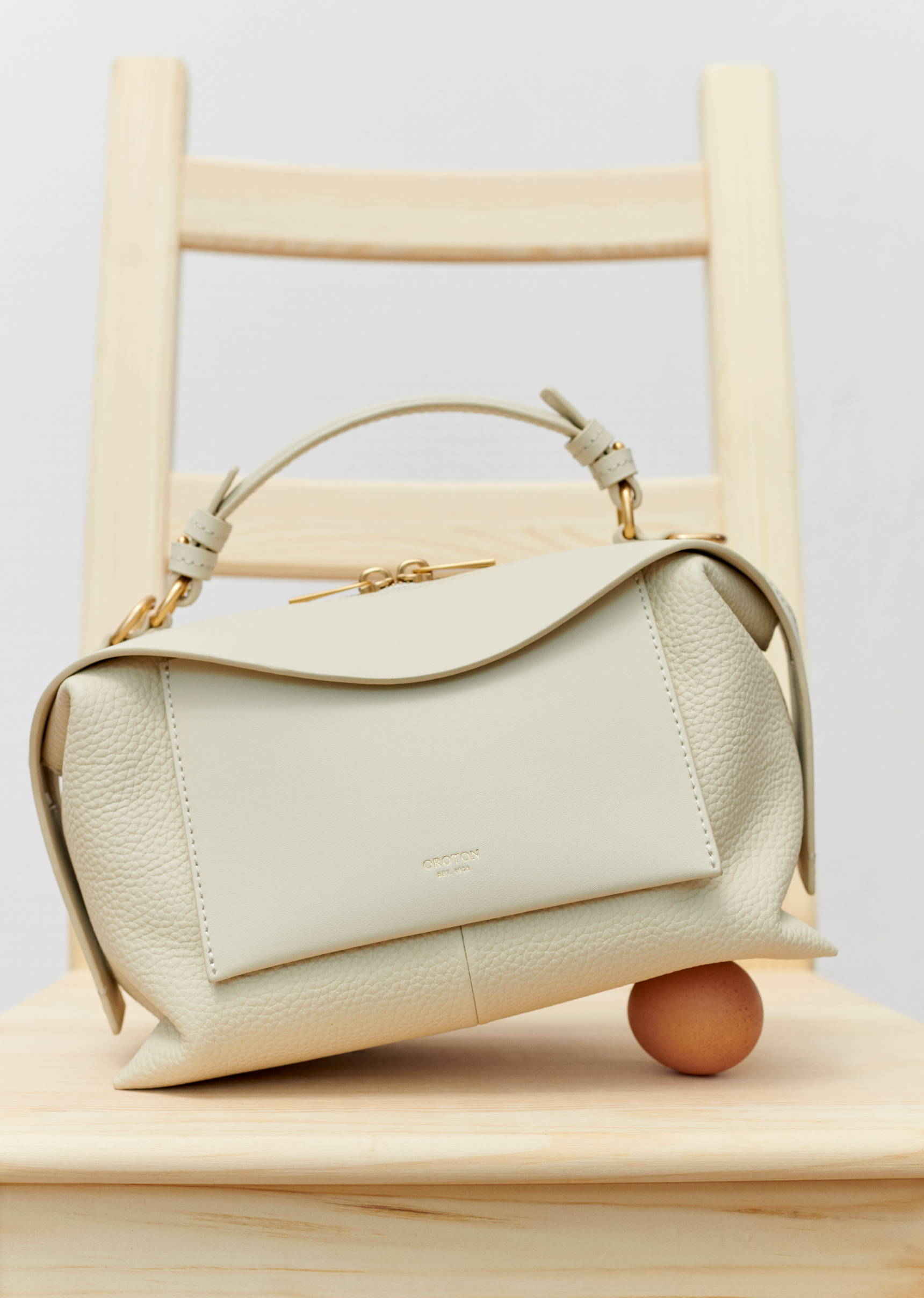 Ivory-colored leather handbag on a light wood chair with an egg underneath it, against a neutral background, in a product photography style.