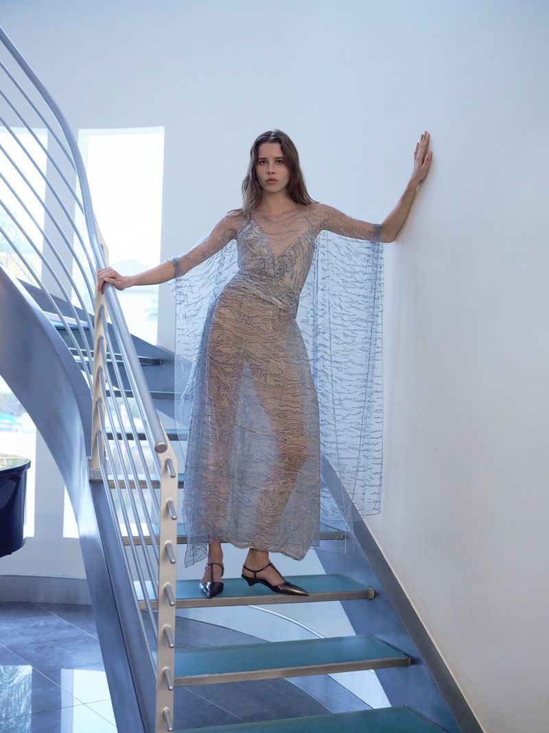 A slender woman models a flowing, translucent dress while posing on modern stairs, bathed in soft, natural light from nearby windows.