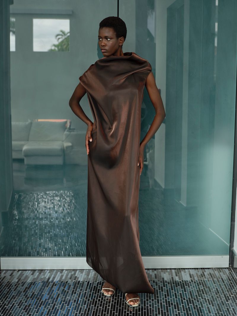 An adult woman poses in a brown dress in front of a glass wall. The model's expression is neutral. Interior setting with tile floor visible.