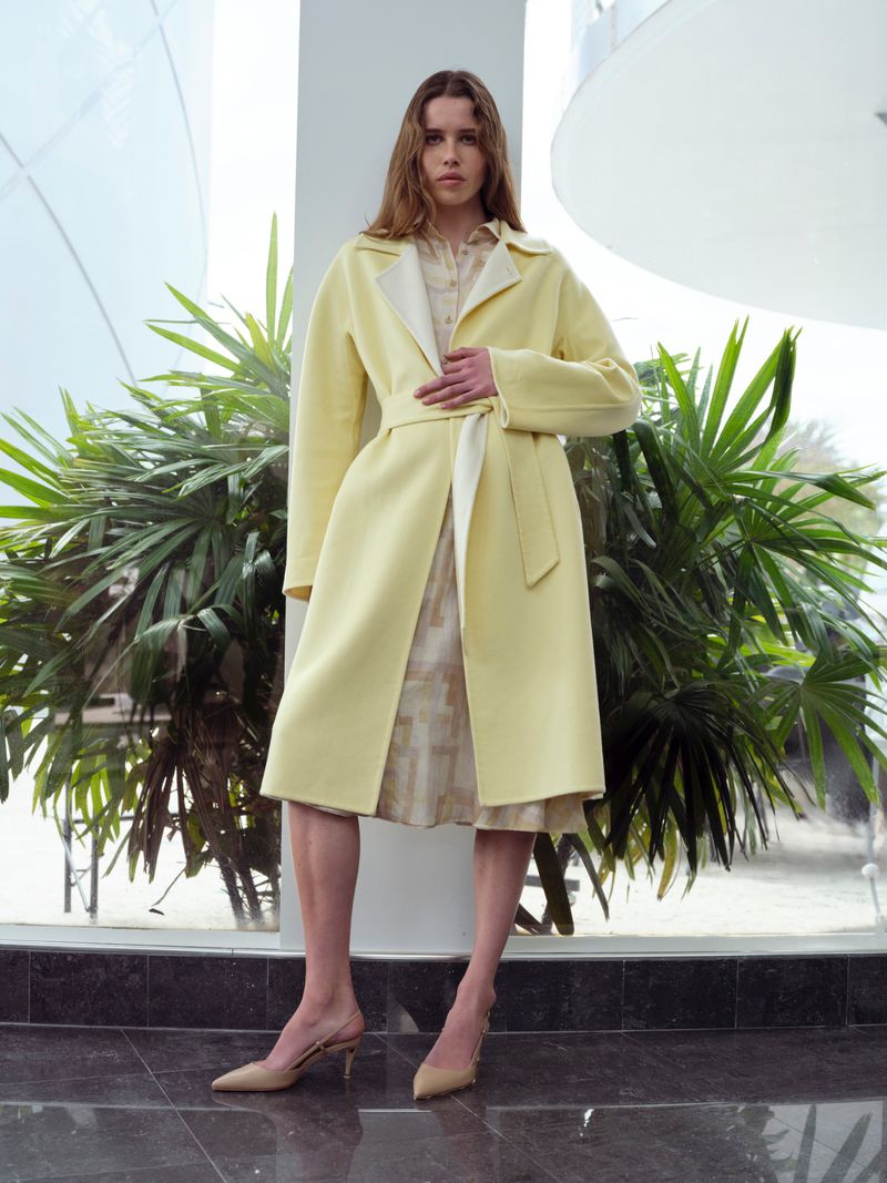 Model in pale yellow belted coat standing indoors near lush tropical plants