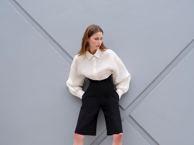 Model wearing a white blouse and black high-waisted shorts leaning against a grey geometric wall